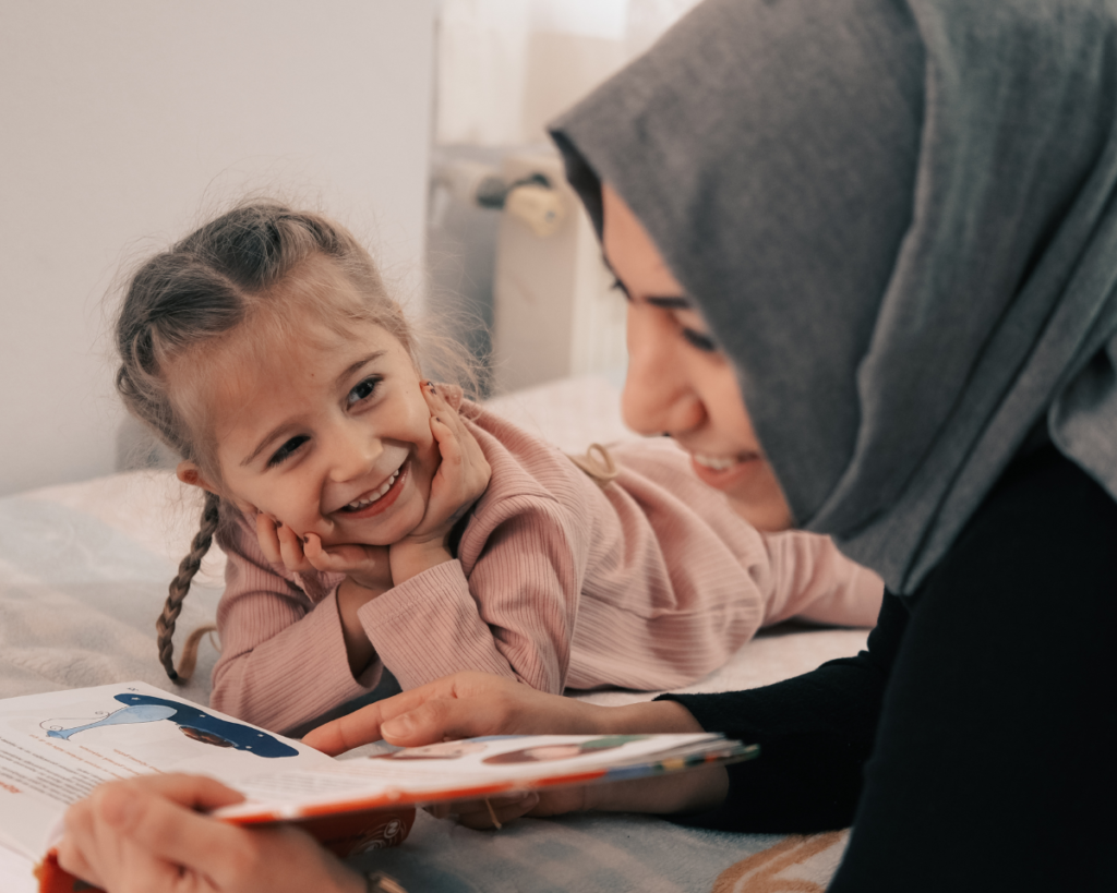 Mother reading a book to daughter, while both are smiling.