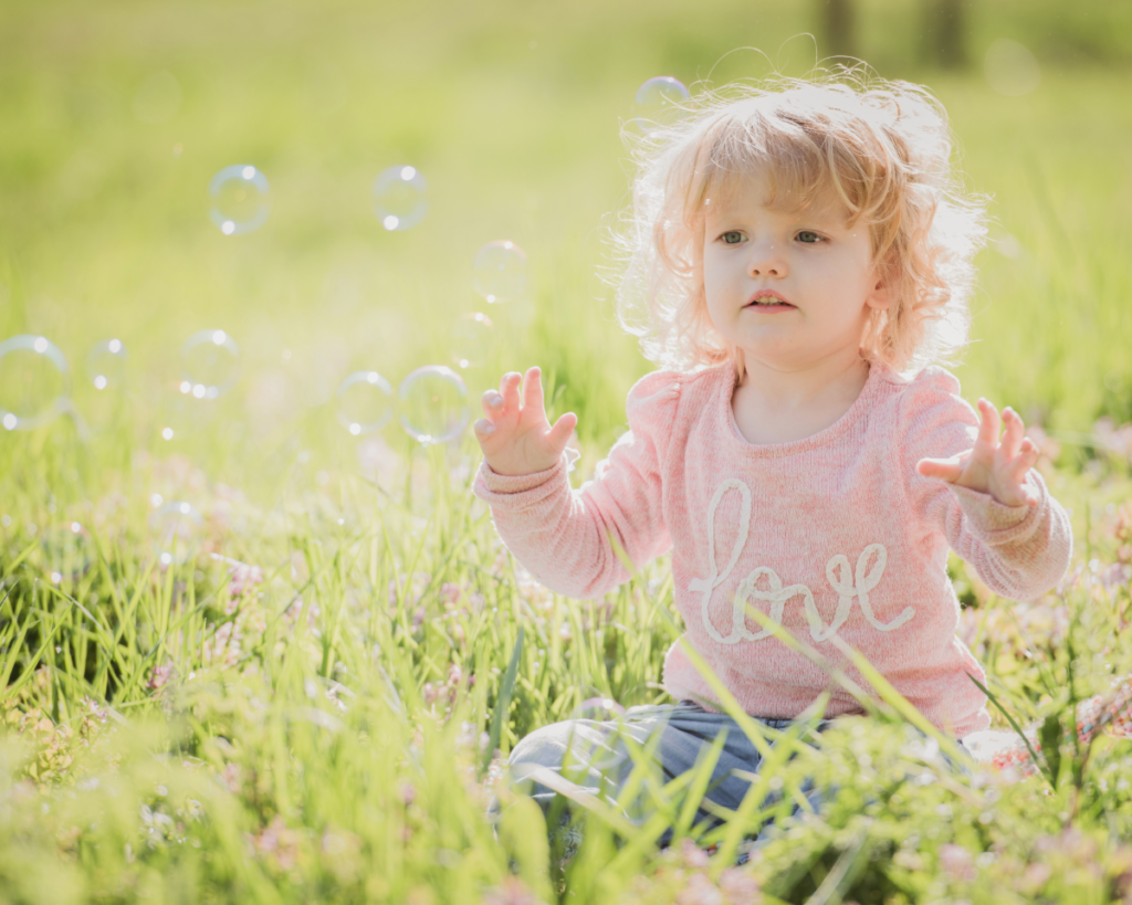 Toddler girl sitting in the grass with bubbles floating around her.