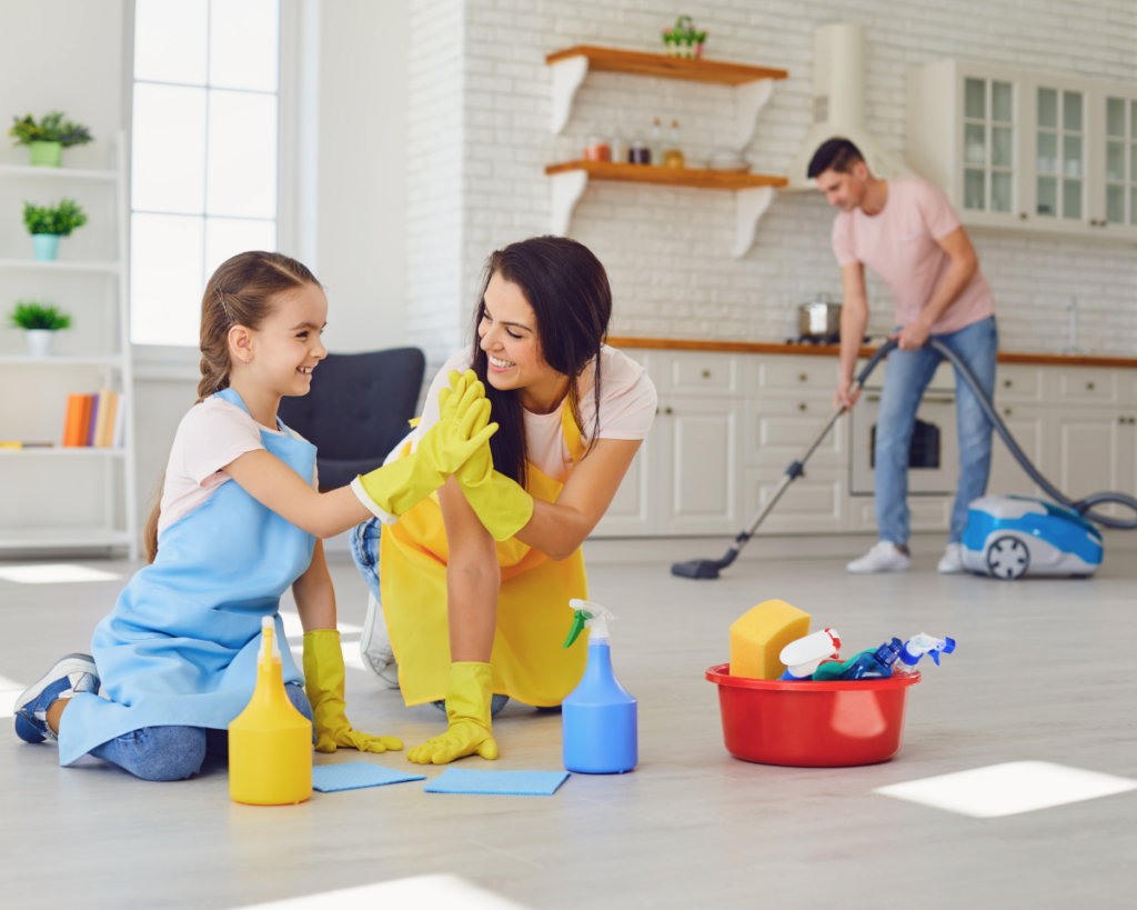 Girl and mom high-five each other while they are scrubbing the floor and dad is vacuuming in the background.