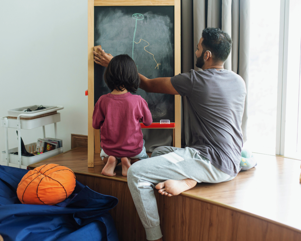 Child and father drawing on chalkboard easel.