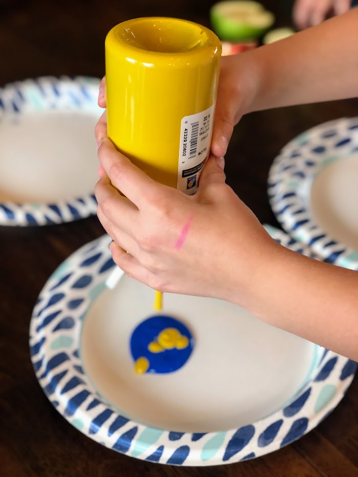 Child's hands squeezing a bottle of yellow paint onto a plate with blue paint.