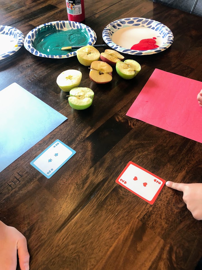 Children's hands with number playing cards, halved apples, papers with trees drawn on them, and plates with paint.