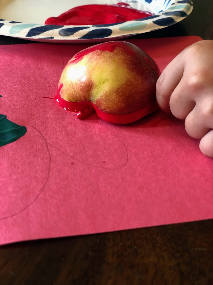 Child stamping a painted apple half on red paper.