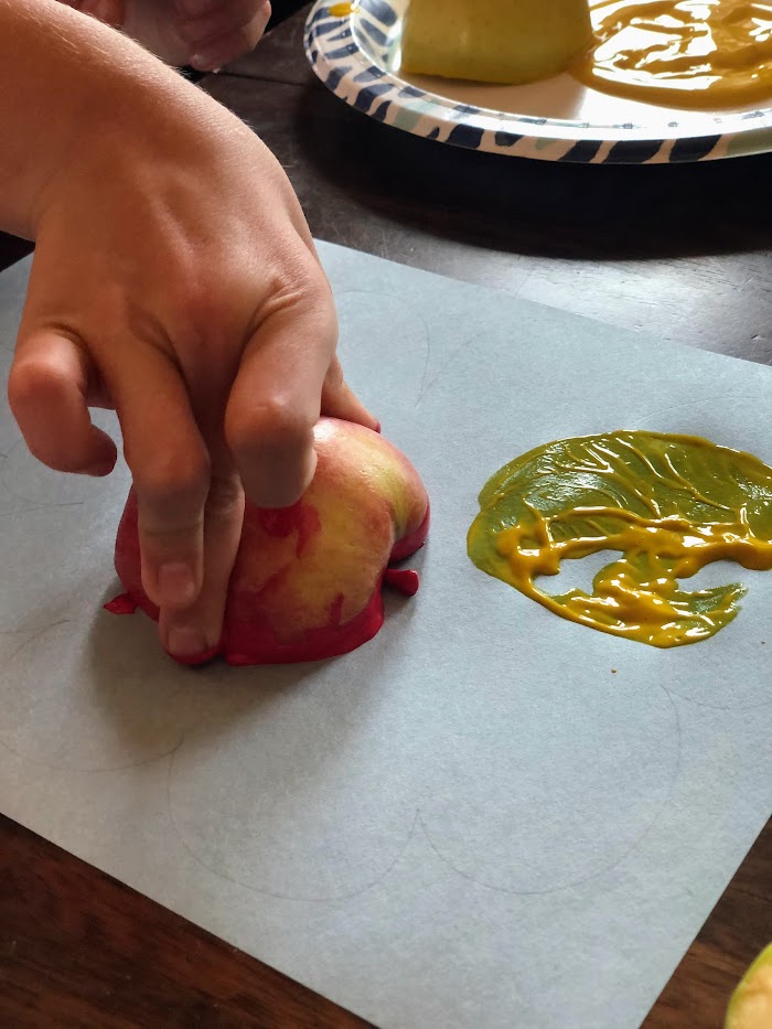 Child's hand stamping a halved apple with red paint on a paper.