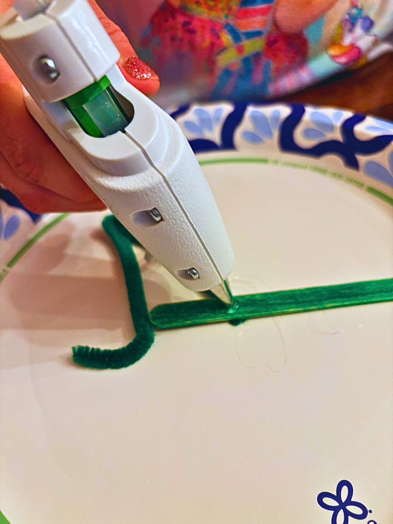 Hot glue gun putting a drop of glue on a green popsicle stick.