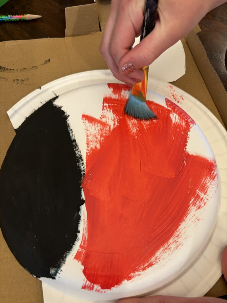Child's hand painting a paper plate red to make a ladybug craft