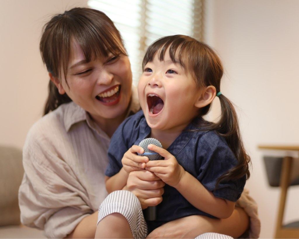 Preschool age girl singing into a microphone while sitting on her mother's lap.