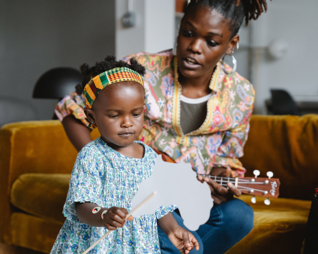 A toddler girl holding a prop for singing, while her mother sings and plays a guitar.