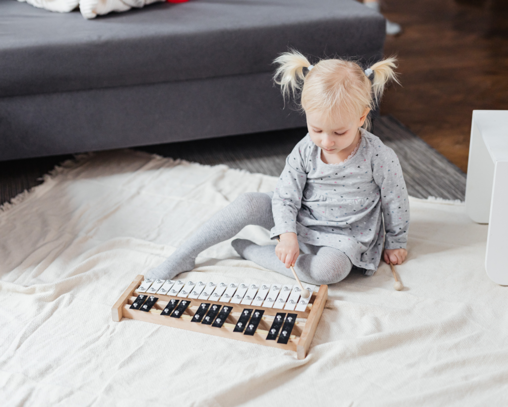 Preschool age blonde girl with pigtails and gray dress. She is sitting on a white blanket playing music on a xylophone.