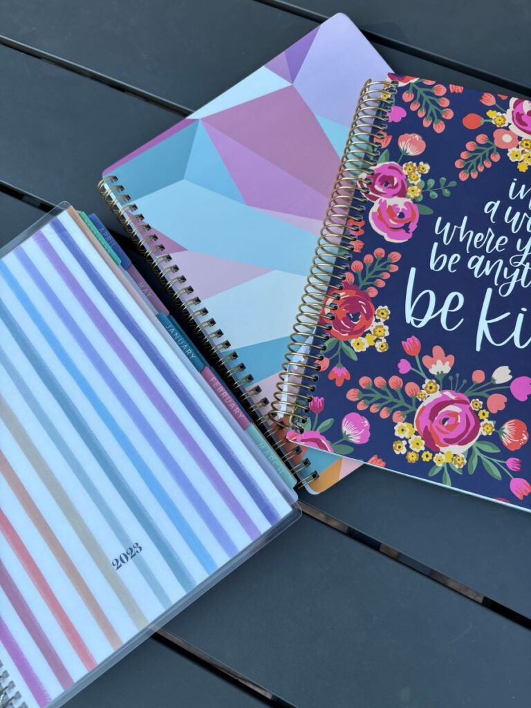 Photo of 3 spiral bound planners. Left planner has vertical stripes in rainbow colors. Middle planner has geometric prism shapes in pastel colors. The planner on the right is navy with pink, red, and yellow flowers.