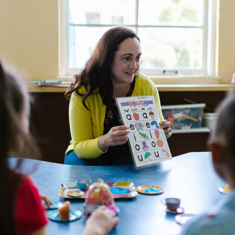 Teaching sitting at a small group table holding a chart with vowels to a group of children.